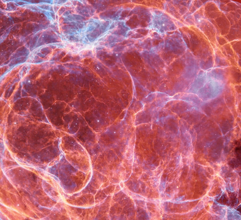 A vibrant image of glowing, colorful nebulae in space, featuring swirling red, orange, and blue filaments of gas and dust against a dark background, resembling intricate cosmic clouds.