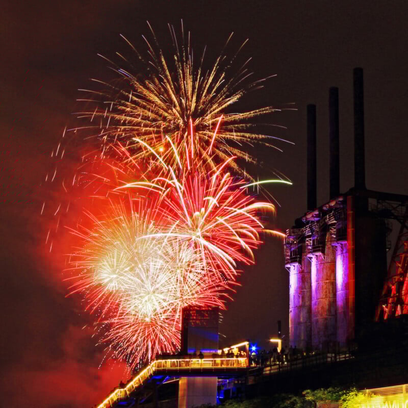 Colorful fireworks burst in the night sky beside an old industrial building with tall smokestacks, illuminated by lights along a walkway below.