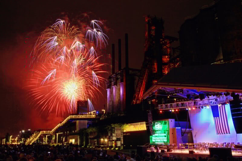 Colorful fireworks light up the night sky above a large outdoor stage with an American flag, an audience, and illuminated industrial structures in the background.