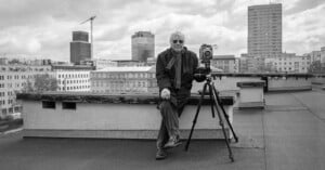 An older man wearing sunglasses sits on a rooftop beside a large camera on a tripod, with a cityscape and tall buildings in the background on a cloudy day.