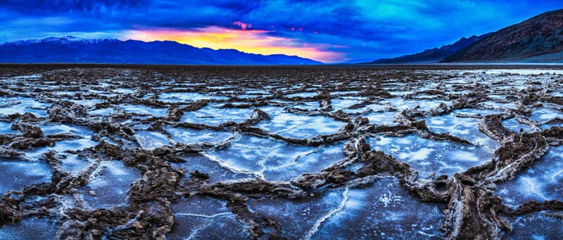 Salt-crusted patterns cover the ground in a desert landscape at sunrise, with mountains in the distance under a vibrant blue and orange sky.
