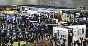 A large, busy convention hall filled with exhibition booths from major camera brands like Sony and Nikon, with many people walking, browsing, and interacting at the event.