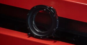 A black metal camera lens mount adapter ring with a "LOCK" label is placed upright against a textured red and black background. The adapter's circular shape and detailed markings are visible.