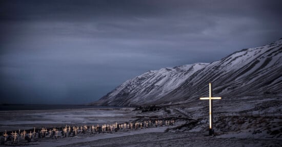 A snowy, coastal graveyard with rows of illuminated crosses at dusk, set against dramatic mountains and a cloudy sky. One large glowing cross stands prominently in the foreground.