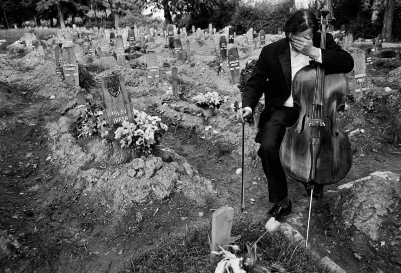 A man in a suit sits on a cello, covering his face with one hand, in a cemetery surrounded by graves and flowers, conveying a sense of grief and mourning.