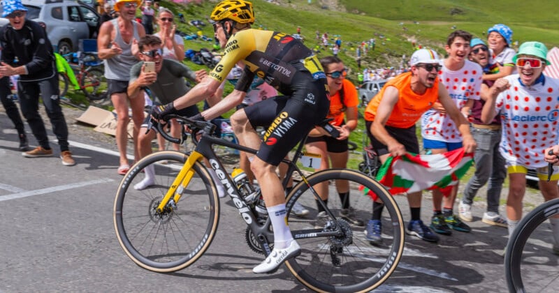 A cyclist in a yellow helmet and racing attire rides uphill, surrounded by cheering fans in colorful outfits and costumes on a mountain road. Fans wave flags and encourage the cyclist enthusiastically.