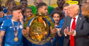 A group of soccer players in blue jerseys celebrate with a large gold trophy, while a man in a suit and red tie stands nearby applauding. The scene is lively, with many players smiling and cheering.