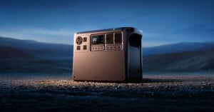 A portable power station with multiple outlets and a digital display sits on a barren, rocky landscape at dusk, with mountains and a cloudy sky in the background.