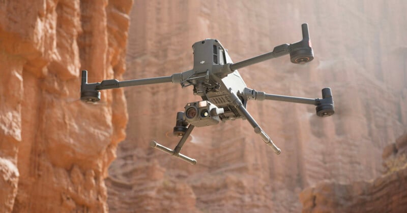 A quadcopter drone equipped with cameras and sensors hovers in front of tall, rugged red rock formations in a canyon, with sunlight streaming through the scene.