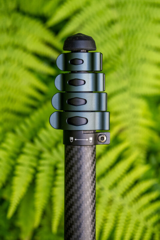 A close-up of a carbon fiber tripod leg with three twist locks, set against a blurred background of green fern leaves.