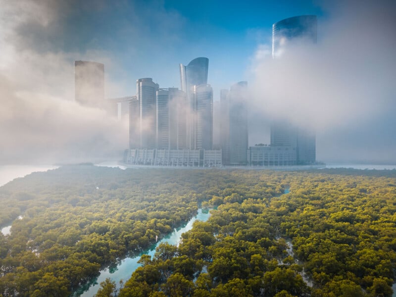 A cluster of modern skyscrapers rises above dense morning fog, with lush green mangroves and winding waterways in the foreground under a clear blue sky.