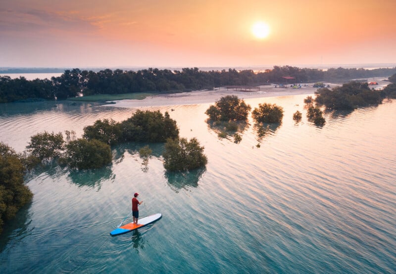 A person on a paddleboard glides across calm water at sunset, surrounded by small clusters of trees and a distant shoreline with vegetation under an orange, hazy sky.