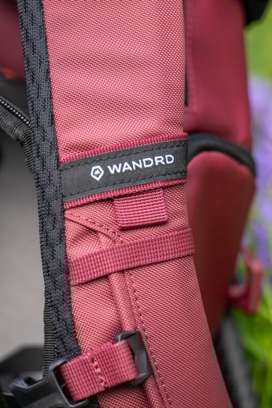 Close-up of a maroon backpack strap with black mesh lining, featuring the "WANDRD" logo in white on a black tag.