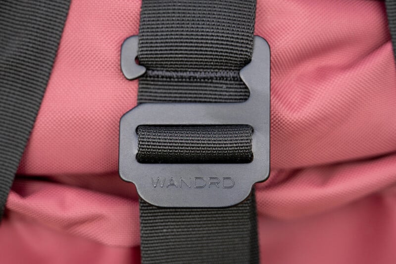 Close-up of a black plastic buckle with the brand "WANDRD" on it, attached to a black strap, against a pink fabric background.