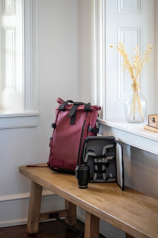 A maroon backpack sits on a wooden bench beside a camera insert, a camera lens, and a glass vase with dried wheat stems, near a sunlit window with white trim.