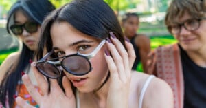A young woman with long nails adjusts her sunglasses and looks at the camera, with two friends in the background, outdoors on a sunny day in a park.