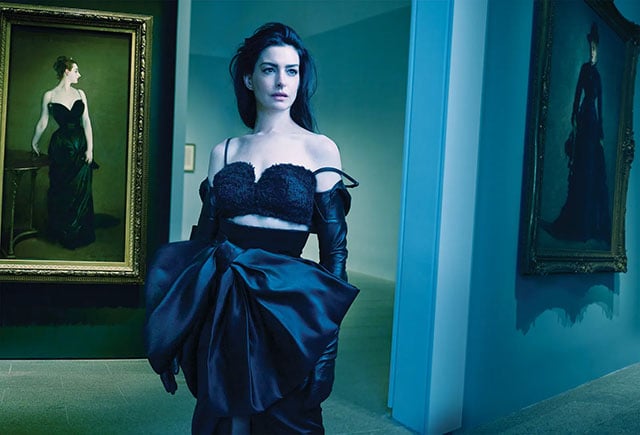 A woman in a glamorous black outfit and gloves stands in an art gallery with teal lighting, next to a large framed portrait of a woman in a black dress.