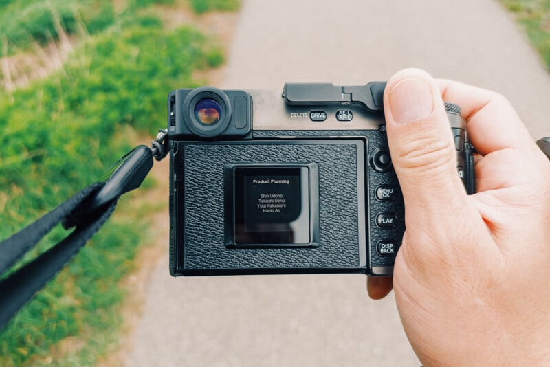 A hand holds a black digital camera outdoors, with the viewfinder screen displaying "Predict Planning." A blurry path and green grass are visible in the background.