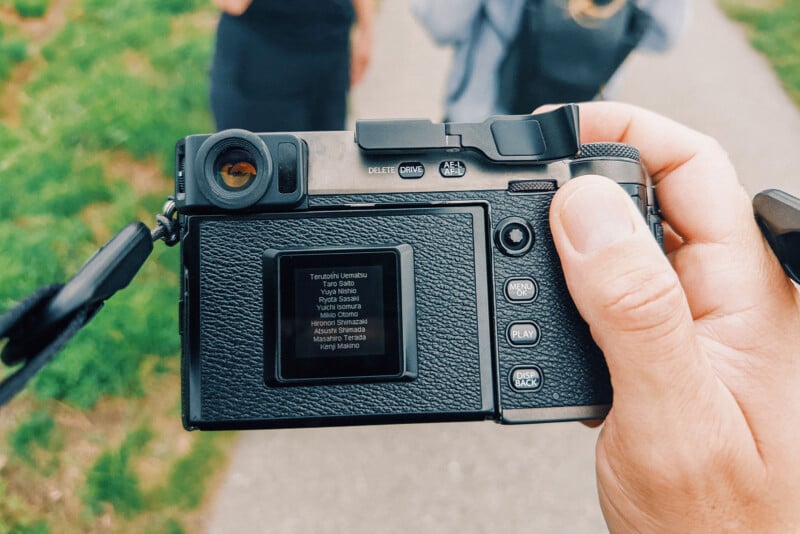 A hand holds a digital camera, showing its rear display screen with a list of names on it. The camera is outdoors on a path, with blurred figures and grass in the background.