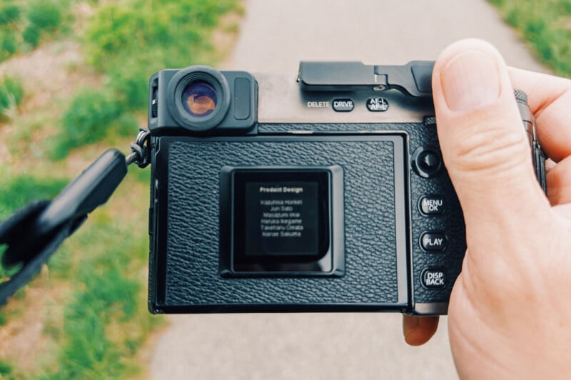 A close-up of a person's hand holding a digital camera outdoors, with the camera's screen displaying a blurry menu or text. The background shows a pathway and green grass.