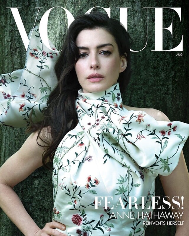 Anne Hathaway poses on the cover of Vogue, wearing a floral, high-neck gown with dramatic ruffles, standing against a textured green background. The word "FEARLESS!" and her name are featured in bold text.