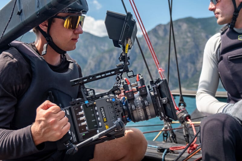 A man wearing sunglasses and a helmet operates a professional video camera on a boat, with mountains and water in the background. Another person in sailing gear is seated nearby.