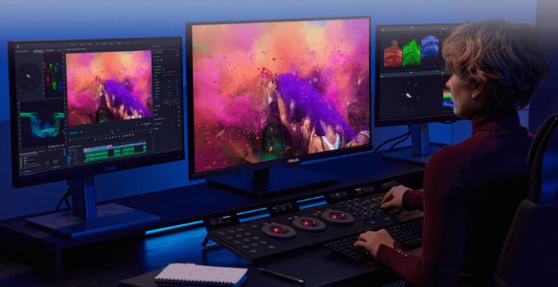 A person uses a color grading console and three monitors displaying vibrant, colorful video editing software and color correction tools in a modern editing suite.