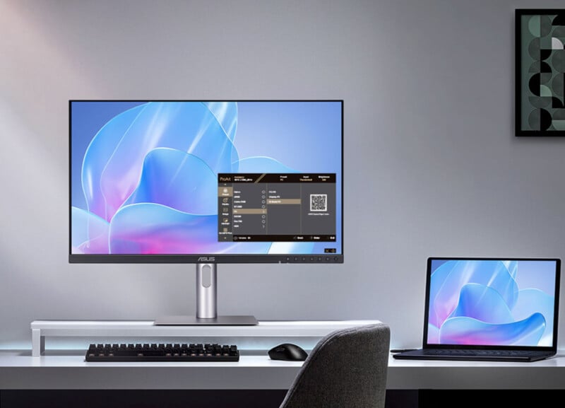 A modern workspace with an ASUS monitor displaying settings, a keyboard, a mouse, and a laptop on a white desk against a gray wall with a framed abstract artwork.