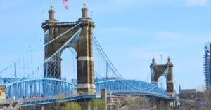 The John A. Roebling Suspension Bridge spans a river, featuring blue cables and two stone towers with gold-topped spires. American flags fly atop each tower, and modern buildings are visible in the background.