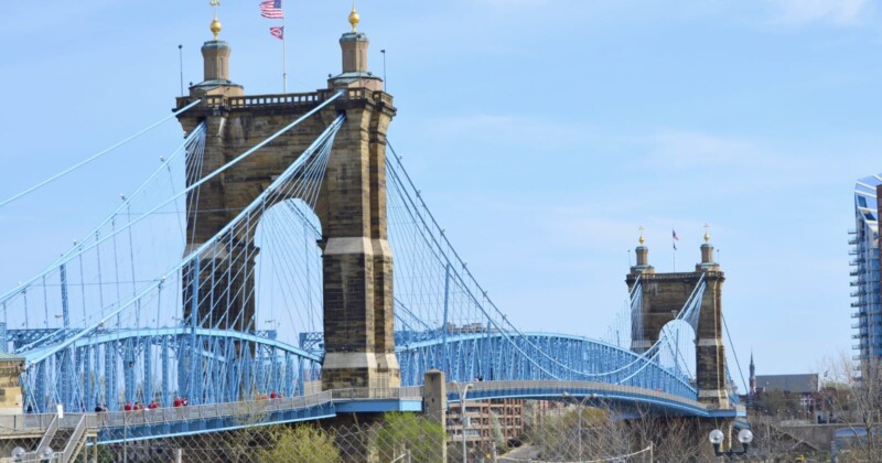 The John A. Roebling Suspension Bridge spans a river, featuring blue cables and two stone towers with gold-topped spires. American flags fly atop each tower, and modern buildings are visible in the background.