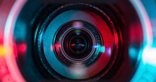Close-up image of a camera lens, with reflections and colorful lighting in shades of red and blue highlighting the lens rings and glass.