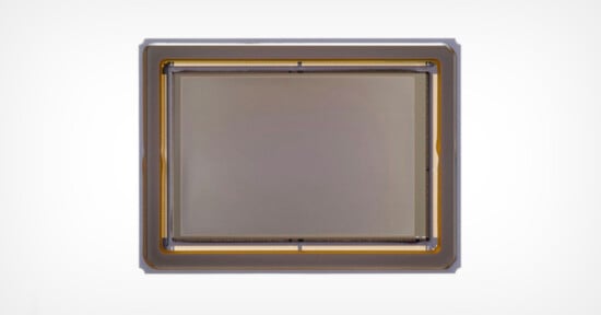 Close-up view of a rectangular camera sensor with a metallic frame and gold wiring visible along the edges, set against a plain white background.