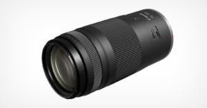 A black Canon RF 100-400mm telephoto camera lens with textured zoom and focus rings, a control switch, and white markings, displayed against a white background.