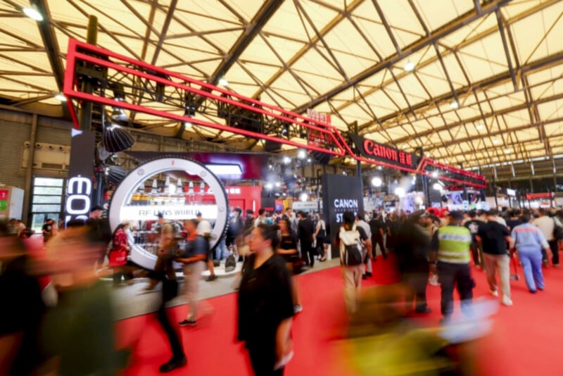 A busy convention or trade show floor with many people walking past a large Canon booth featuring prominent signage and bright lights under an industrial ceiling. The scene is lively and bustling with activity.