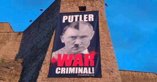 A large banner hangs on a stone wall, featuring a photoshopped image combining Vladimir Putin’s and Adolf Hitler’s faces, with the words "PUTLER WAR CRIMINAL!" in bold letters below.