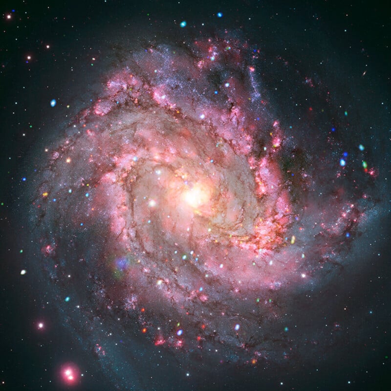 A bright, colorful spiral galaxy with glowing pink, blue, and white regions, surrounded by numerous stars set against a dark space background.