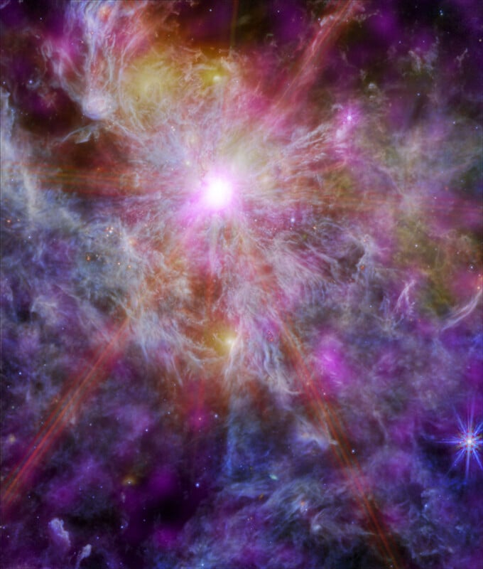 Colorful space image showing a bright, glowing star at the center, surrounded by swirling clouds of gas and dust in shades of purple, blue, and gold, with a smaller bright star at the bottom right.