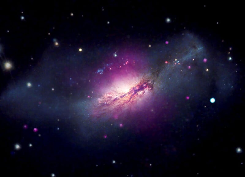 A colorful galaxy glows in deep space, with a bright pink and purple center surrounded by clouds of blue and white dust, against a black background dotted with stars.