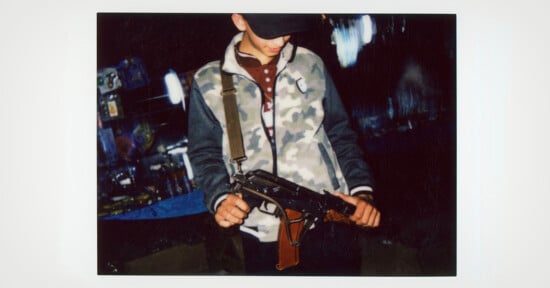A young person wearing a cap and camouflage jacket looks down while holding a toy or replica rifle, standing in a dimly lit area with blurred lights in the background.