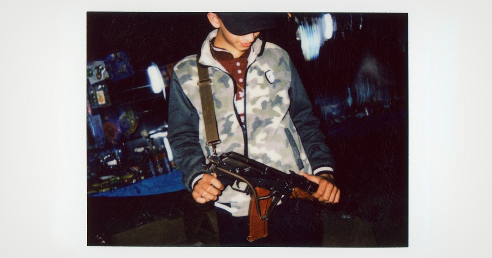 A young person wearing a cap and camouflage jacket looks down while holding a toy or replica rifle, standing in a dimly lit area with blurred lights in the background.