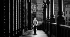 A person wearing a hooded jacket walks alone on a bridge, framed by tall columns and lamps, in a black-and-white urban setting.
