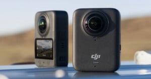 Two gray DJI 360-degree action cameras are positioned on a reflective surface outdoors, one showing its front lens and logo, the other displaying its rear screen with a landscape image.