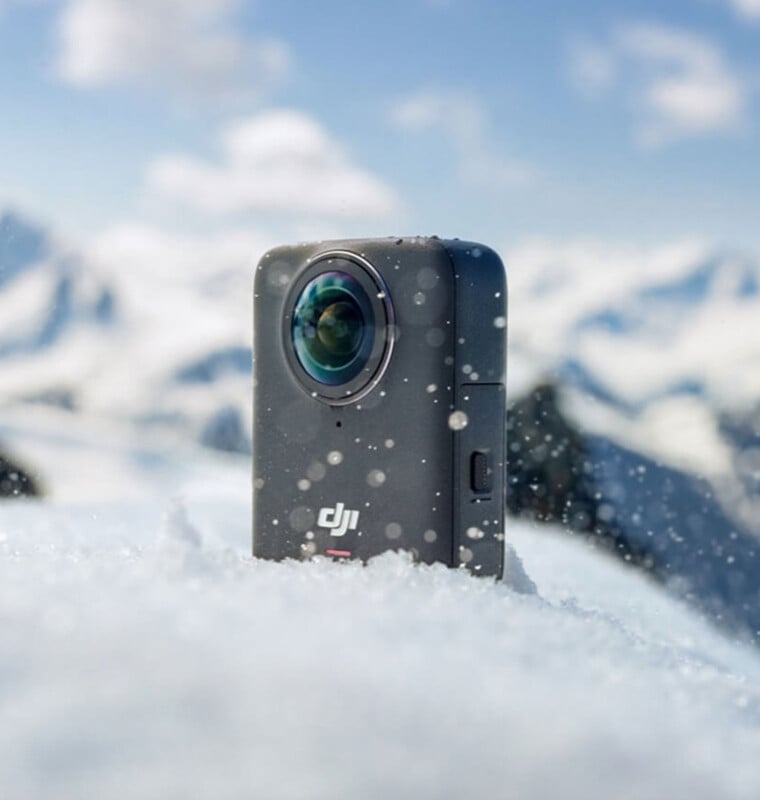 A DJI action camera sits partially buried in snow, with snowflakes in the air and blurred, snowy mountains in the background under a blue sky.