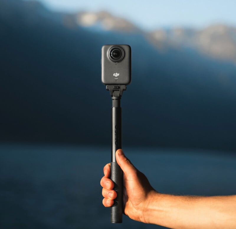 A hand holds a DJI 360 camera mounted on a black selfie stick outdoors, with a blurred background of water and mountains.
