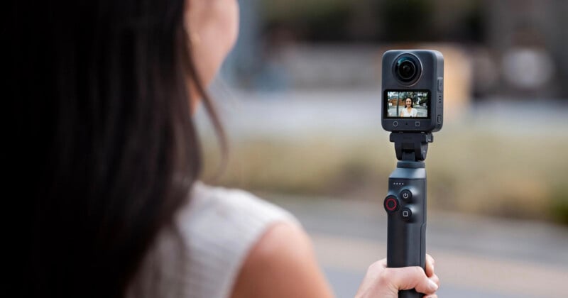 A woman holding a handheld 360-degree camera on a gimbal, capturing an outdoor scene visible on the camera’s screen. The image is focused on the device, with the background blurred.