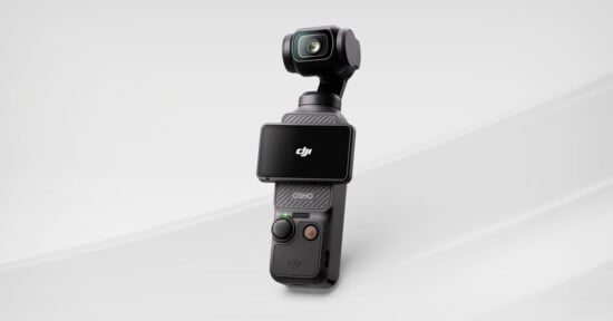 A compact black handheld camera with a built-in gimbal and a flip-out screen displaying the DJI logo, set against a light gray background.