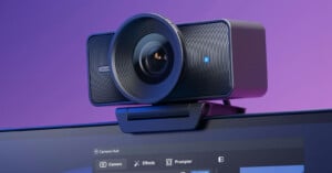 A close-up of a modern webcam with a large lens, mounted on top of a computer monitor. The screen below shows a user interface with camera settings. The background is purple.