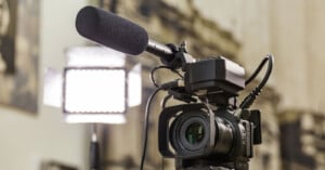 A professional video camera with a large microphone and light panel is set up indoors, ready for filming. The background is out of focus, emphasizing the camera equipment.