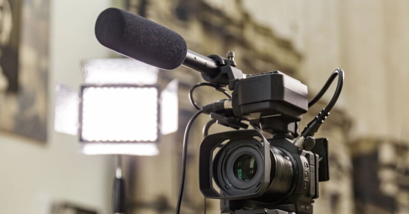 A professional video camera with a large microphone and light panel is set up indoors, ready for filming. The background is out of focus, emphasizing the camera equipment.