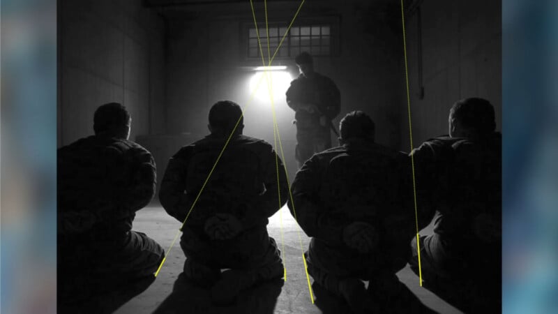 Four people sit on their knees with hands behind their backs, facing a standing figure in military attire, illuminated by light from a barred window in a dark, shadowy room.